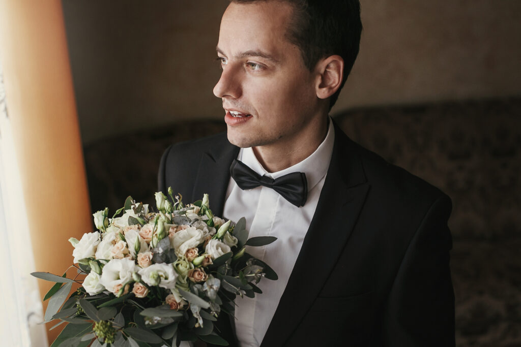 Groom in a black tuxedo holding a bouquet of white and blush roses