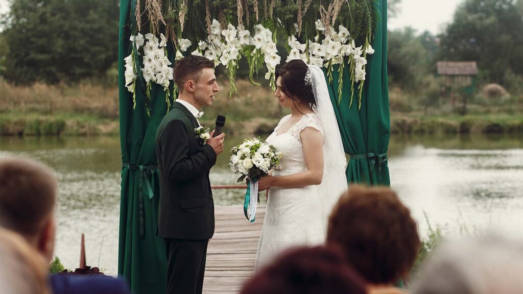Groom reading vows to bride under a floral arch at an outdoor lakeside ceremony